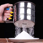 Metal sifter being used to sift flour into a bowl with a dark background