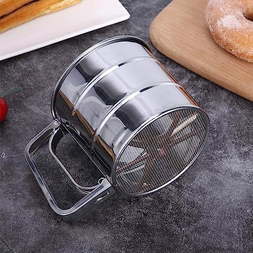 Metal flour sifter on a gray surface with pastries in the background