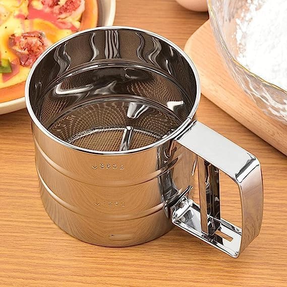Metal sifter on a wooden surface with food items in the background